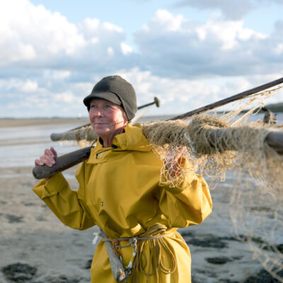 Tessa Bunney - Haaf netting - Sunderland Point (Royaume-Uni), 2018 - Taken in 2018, Margaret Owen was the only woman in the UK licensed to use a ‘haaf net’ for catching salmon and sea trout. For 27 years she has been standing waist deep in the river Lune, 4 hours a day twice a day during the three-month salmon fishing season. “Salmon run upstream on the filling tide and turn back when they meet the fresh water of the river and you stand in the run to catch them”. Now salmon are an endangered species, and from 2019 she was told that EU regulations mean she must discard any salmon she catches. She is allowed to keep the less lucrative sea trout. The image is taken in Sunderland Point which is unique in Britain as being the only community to be on the mainland and yet dependent upon tidal access - via a 1.5 mile long single-track road crossing a tidal marsh.